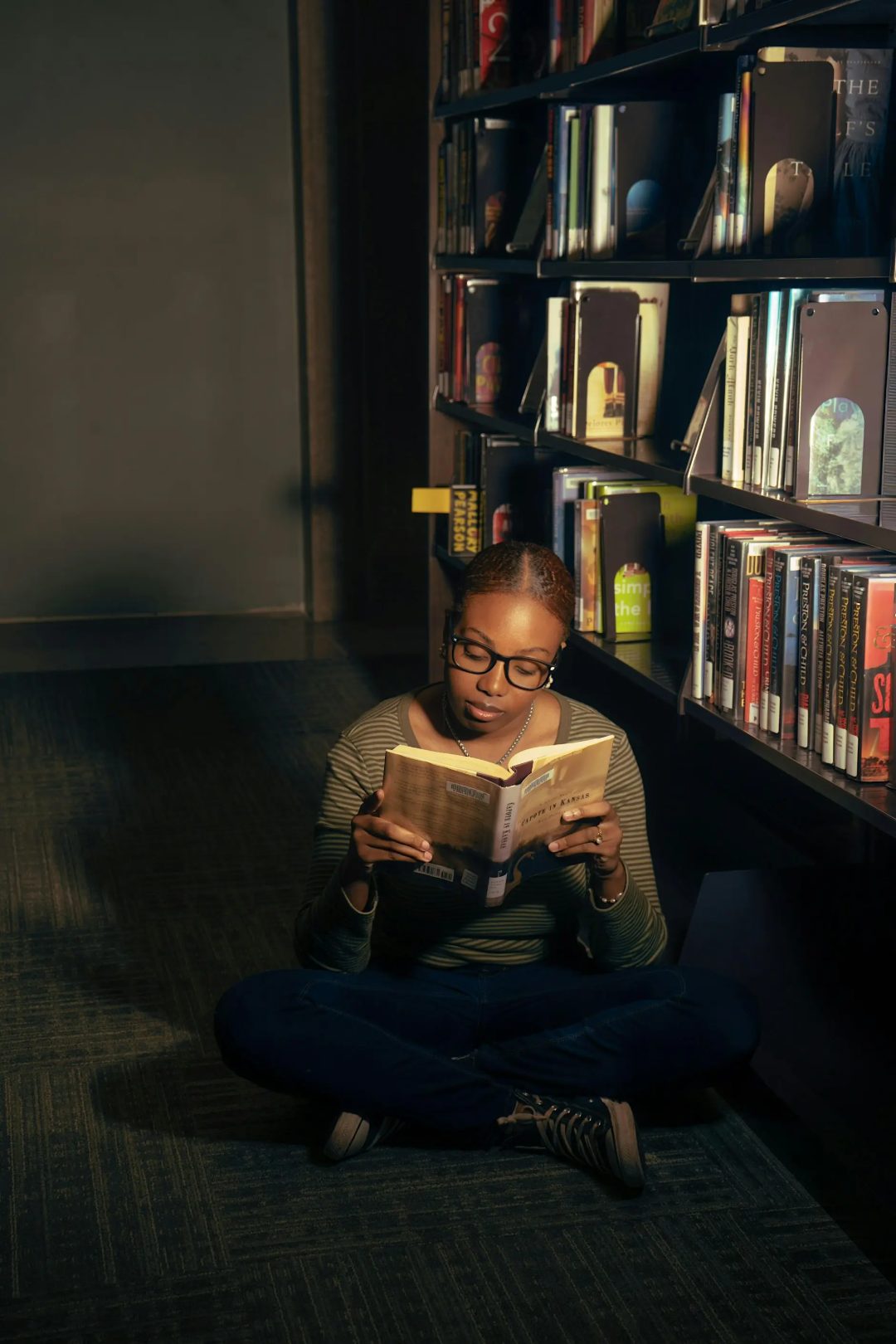 A woman sits with dim lighting reading a book in a library